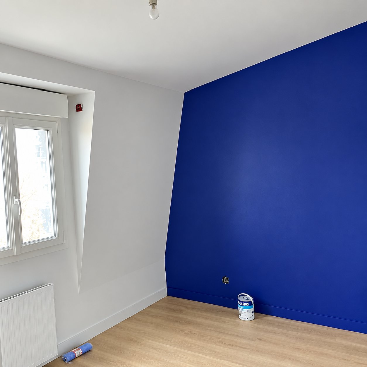 Chambre fraîchement repeinte avec un mur bleu intense, des murs blancs, une fenêtre, un radiateur, un parquet clair et un pot de peinture au sol.