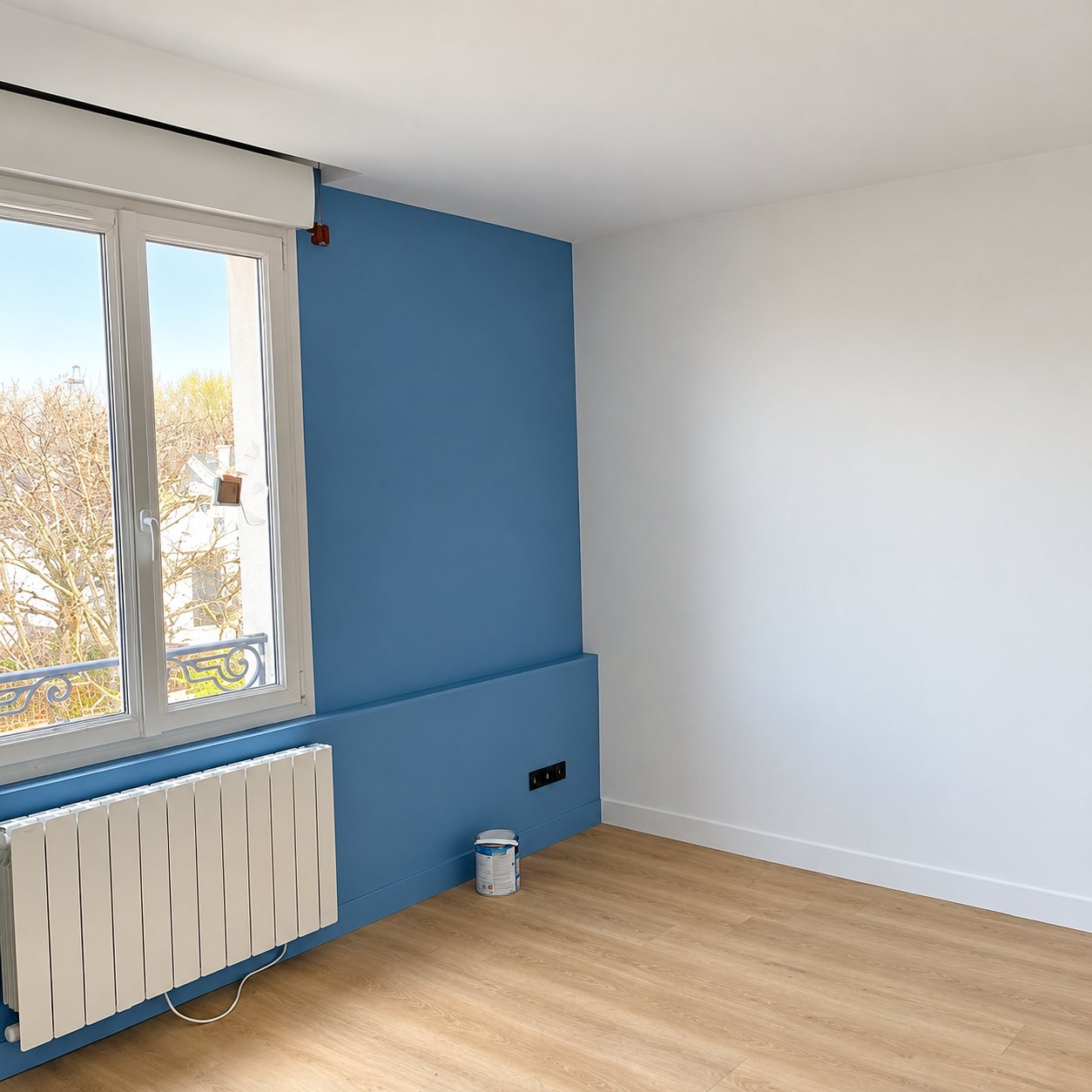 Pièce vide fraîchement rénovée avec un mur peint en bleu, une fenêtre blanche, un radiateur et un parquet clair.