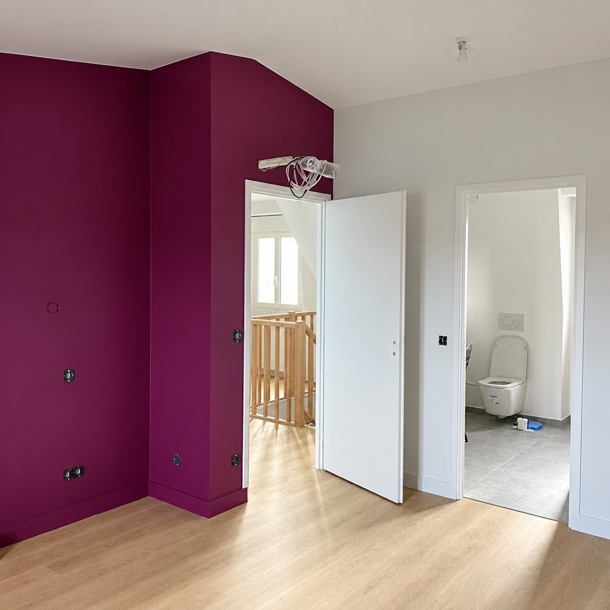 Pièce rénovée aux murs prune et blancs avec parquet clair, porte blanche ouverte donnant sur un escalier en bois et des toilettes suspendues.