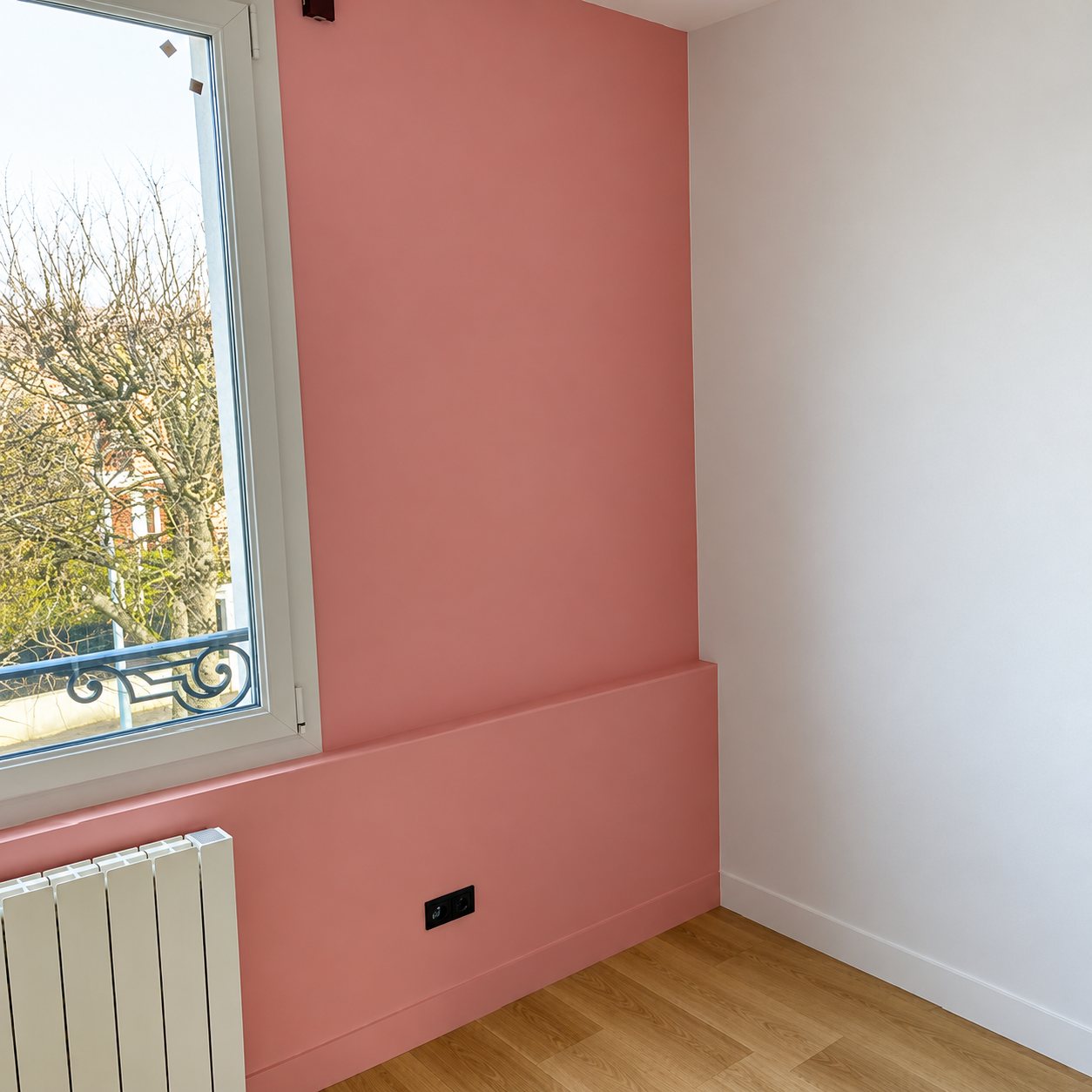 Pièce fraîchement rénovée avec un mur peint en rose, un mur blanc, une fenêtre donnant sur des arbres, un radiateur blanc et un parquet clair.