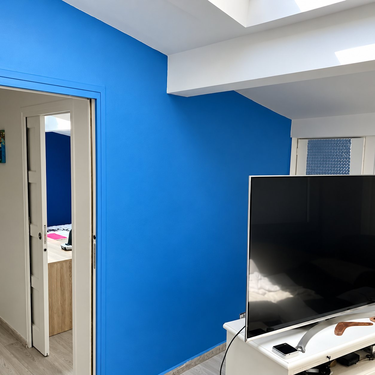 Pièce rénovée aux murs peints en bleu vif, avec une télévision posée sur un meuble blanc et une porte coulissante ouverte sur une autre pièce.